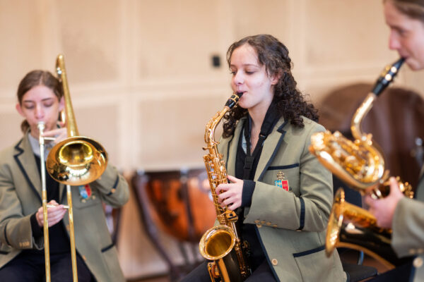 Students in uniforms play saxophones and a trombone in a music room practicing together