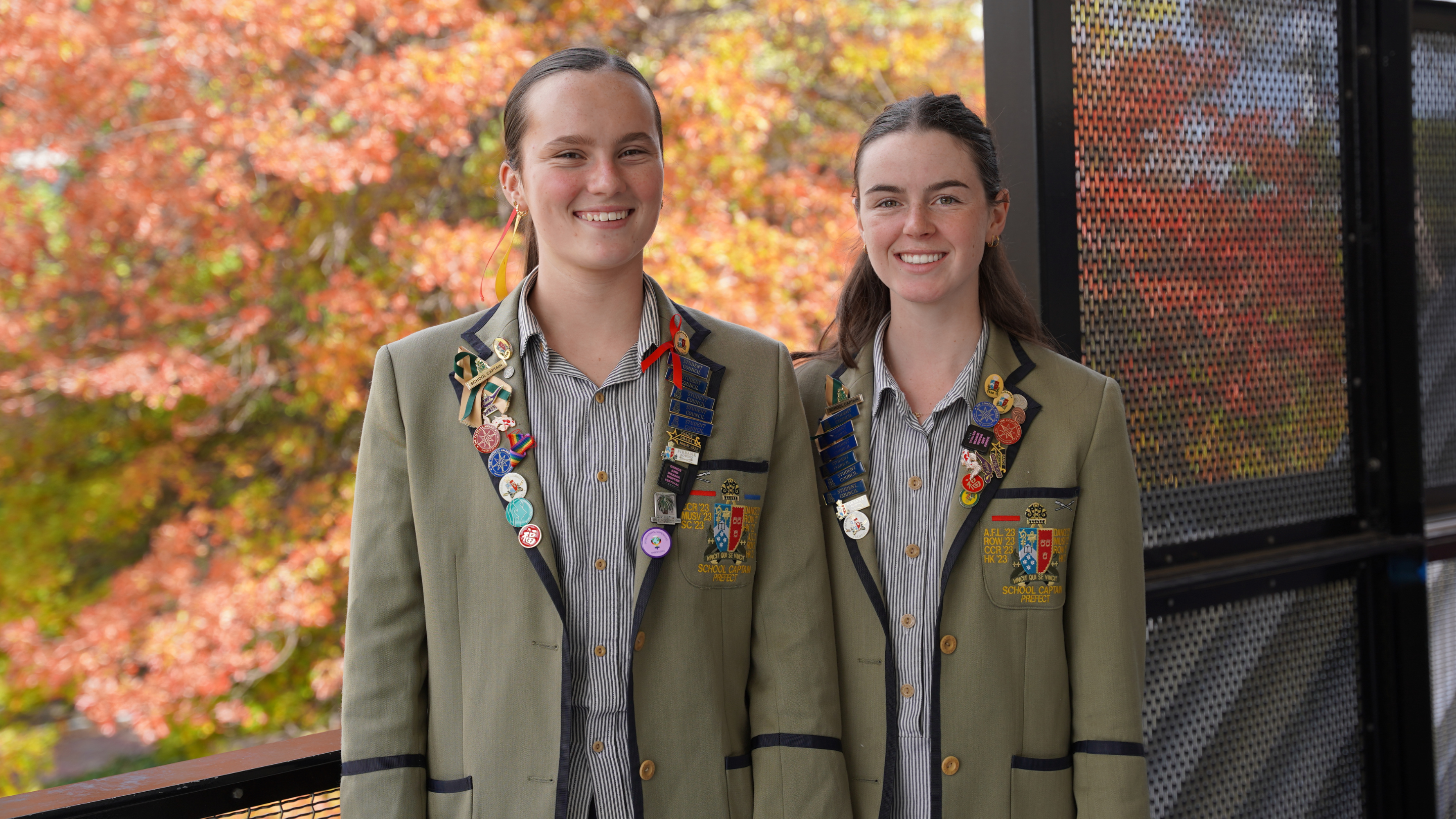 Two students in decorated school blazers smiling outdoors with autumn leaves.