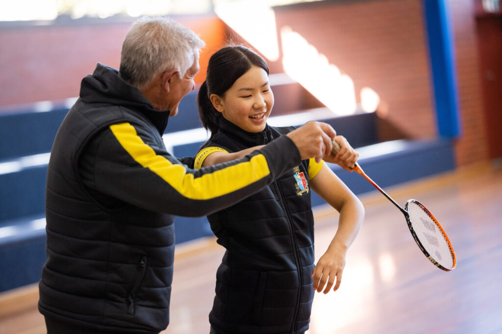 Coach assisting a smiling student with badminton technique in a gymnasium setting