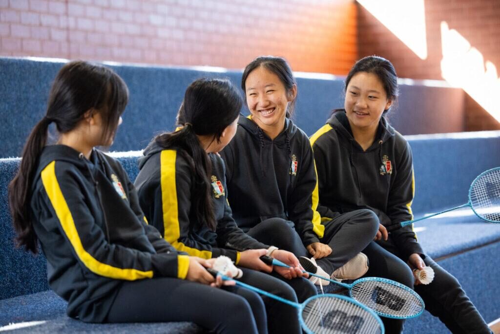 Group of students in sports uniforms sitting on bleachers, holding badminton rackets.