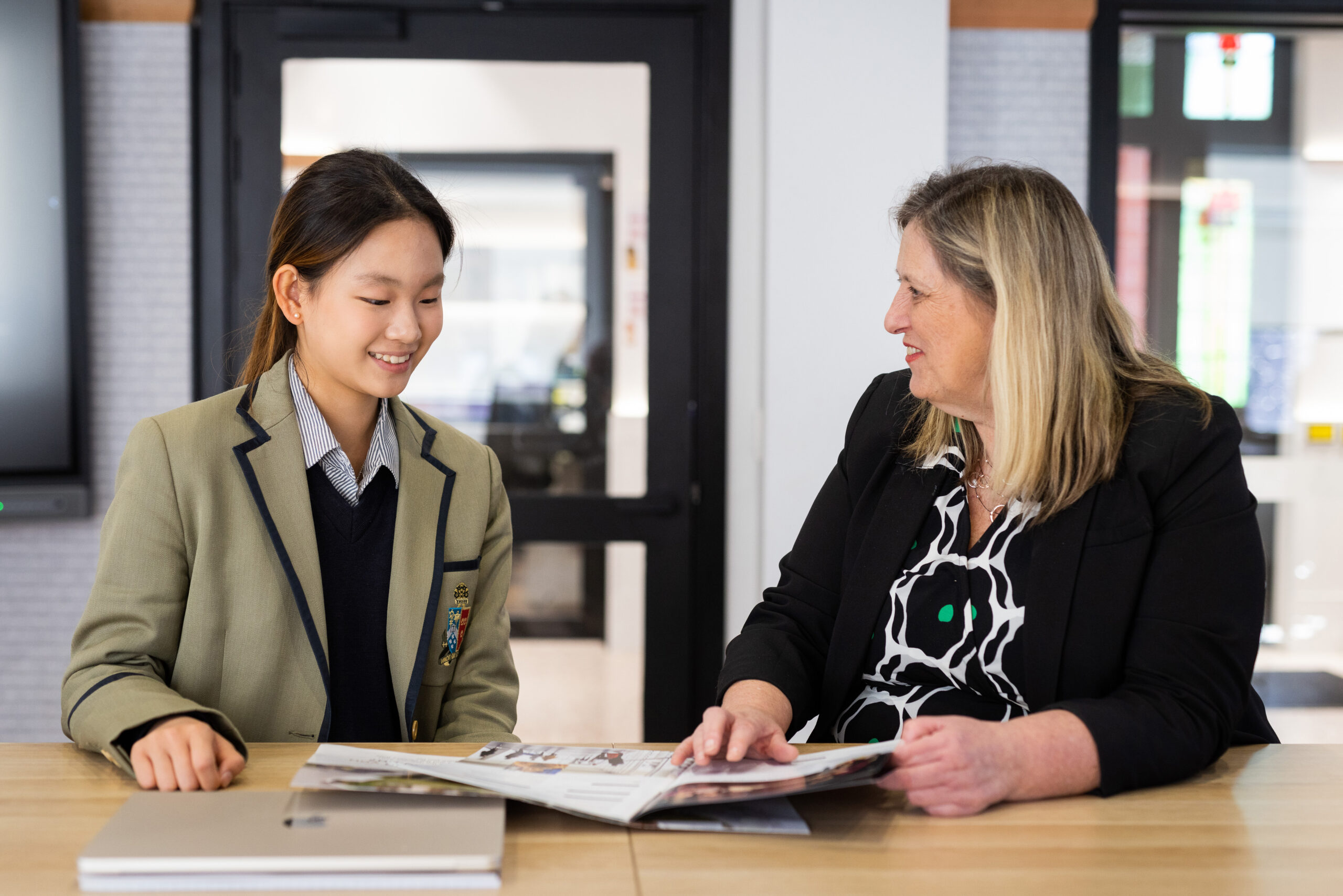 Student in uniform and teacher discussing a book at a table in a classroom setting