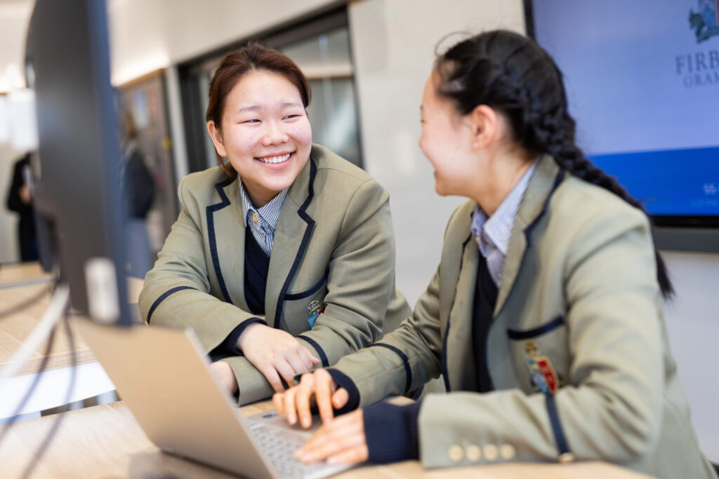 Two students in uniforms smiling and working together on a laptop in a classroom.