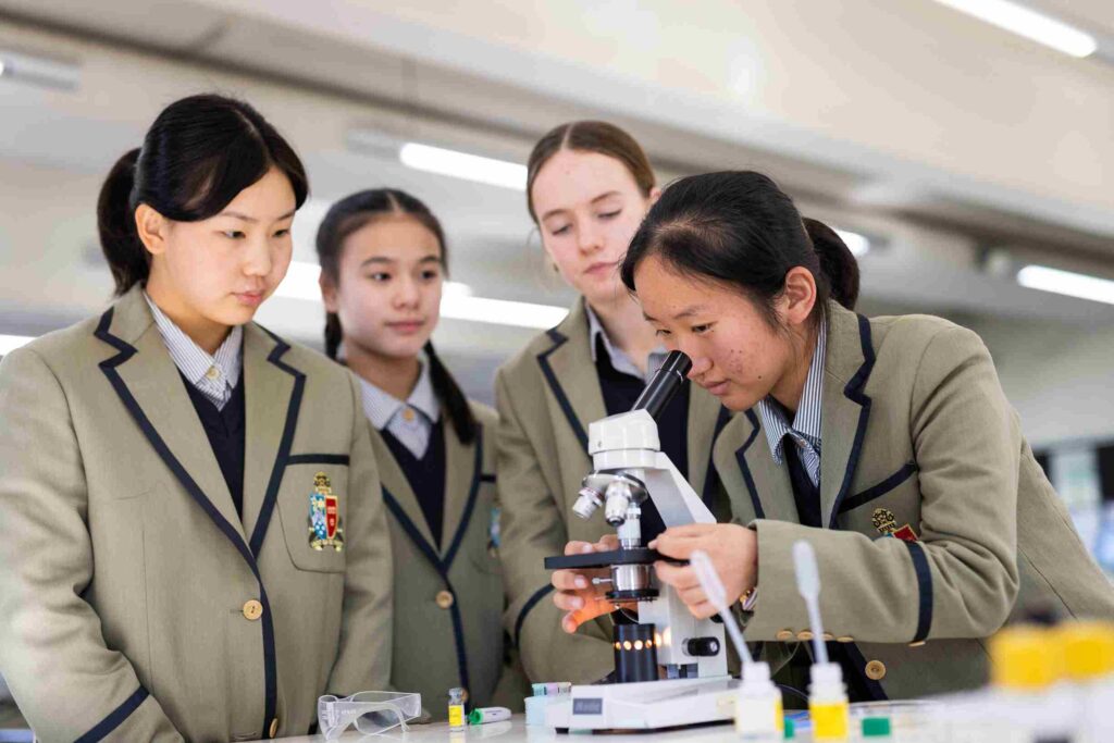 Students in uniforms observing a peer using a microscope in a science lab setting.