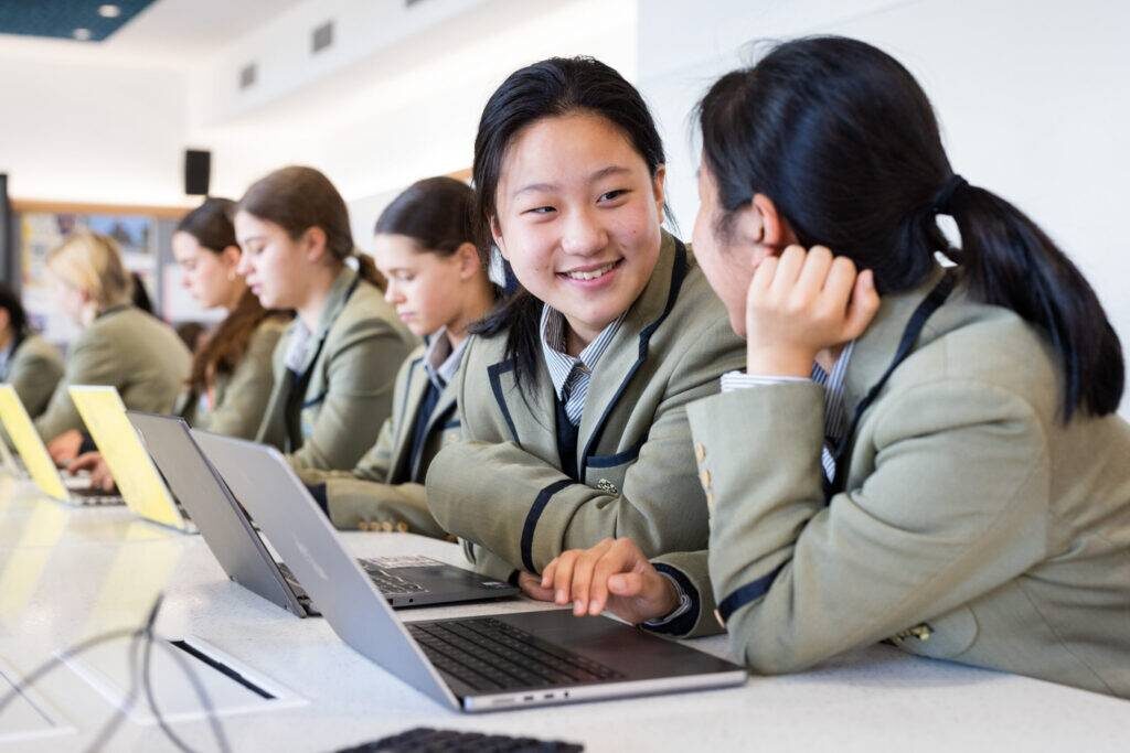 Students in uniforms working on laptops, two girls smiling and chatting together.